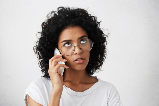 Close-up portrait of bored good-looking black mixed race woman with Afro hairstyle wearing eyeglasses holding cell phone talking with friend looking up having pensive expression. People, communication