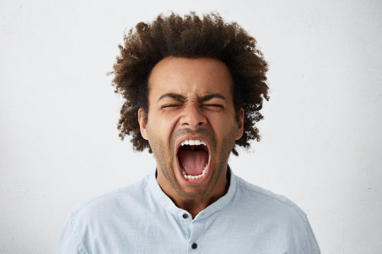 Close-up of Afro American man with bushy hair and pure skin having closed eyes and wide opened mouth screaming with great fear and anger. Furious dark-skinned male with curly hair shouting loudly