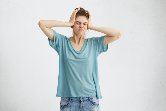 Portrait of stressful fair-haired woman in casual clothes having headache holding hands on her head frowning her face having displeased look. Tired woman having some problems looking stressed