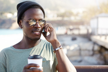 Attractive male with black skin wearing sunglasses calling his girlfriend from abroad to tell about his trip holding takeaway coffee isolated over street background. Phone conversation and rest