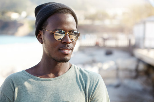 Atttractive young hipster African male in casual shirt , black hat and shades standing against city background. Tourist in trendy wear looking into distance pensively deciding what to explore next