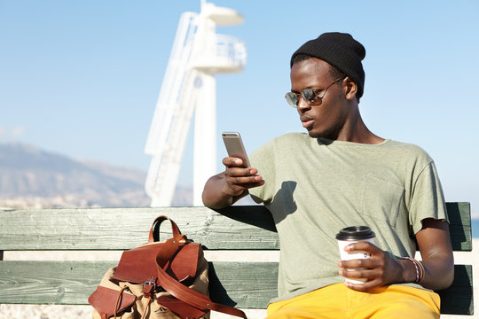Young attractive dark-skinned male wearing black hat, casual T-shirt and yellow trousers sitting at bench near marine landscape reading news online, browsing pictures and drinking delicious coffee