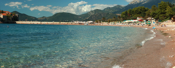 A horizontal panorama overlooking the sea bay with a beach and a lot of people having a rest. Clear clear water, mountains and a cloudy sky in the distance
