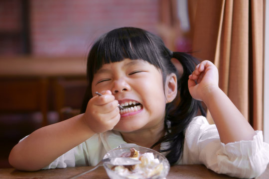 Asian Children Cute Or Kid Girl Enjoy Eating Banana Split Ice Cream And Teeth Smile Or Hypersensitive Teeth On The Wood Table And Window Side With A Curtain In The Restaurant Or Cafe