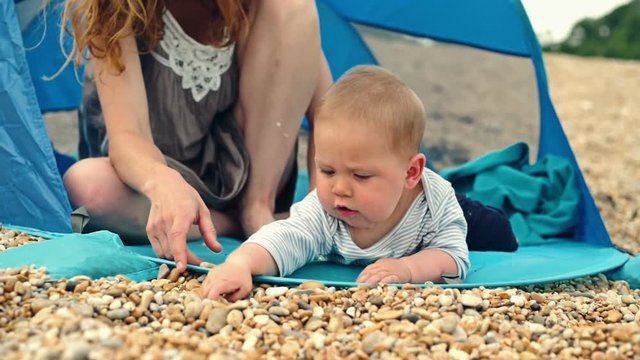 Mother with baby sitting in a tent on the beach. The baby is trying to eat rocks and starts crying when his mother won't let him.