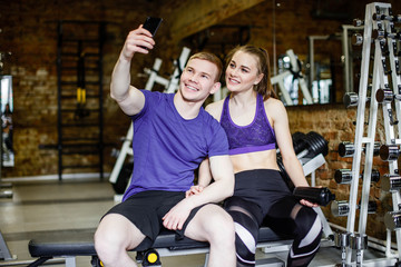 Beautiful happy couple in sports clothes is making selfie using a smart phone and smiling while resting after workout