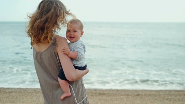 A young her s standing on the beach with her little baby. The baby turns and smiles