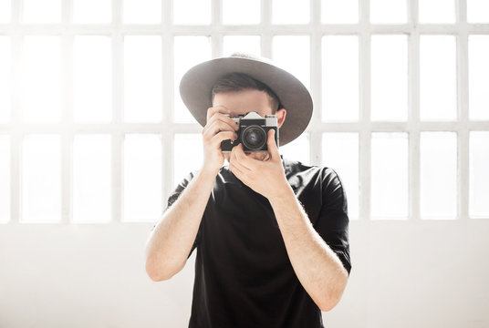 Young handsome photographer in grey hat and black t-shirt takes photos with old film camera in white sunlit room