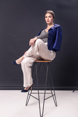 one young Caucasian woman 20s, 20-29 years, fashion model sitting bar stool, posing, studio, white background, fashionable blue jacket, horizontal stripe shirt, pants, high heel shoes