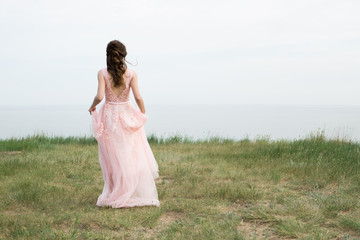 Fototapeta na wymiar Bride in a long pink wedding dress is standing on the seashore. Dress with an open back. Back view.