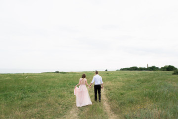 Bride and groom walk around holding their hands on the coast.  On the bride is a long pink wedding dress with lace and an open back.