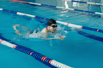 Young woman in goggles and cap swimming breaststroke stroke style in the blue water indoor race pool - obrazy, fototapety, plakaty