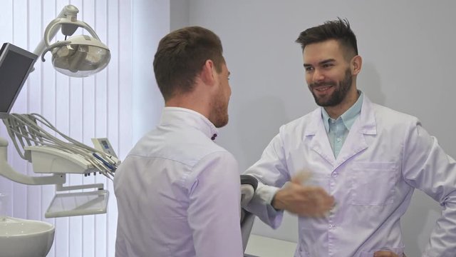 Young bearded dentist appointing patient at the office. Handsome brunette dental specialist shaking hands to his client. Attractive caucasian man meeting his doctor at the clinic