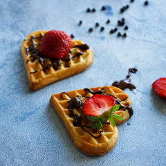 Belgian waffles in the shape of hearts with fresh strawberries and chocolate topping on blue table. Homemade healthy breakfast, selective focus