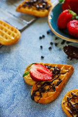 Belgian waffles in the shape of hearts with fresh strawberries and chocolate topping on blue table. Homemade healthy breakfast, selective focus
