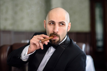 A brutal man in a dress coat smokes a cigar.