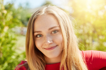 Naklejka premium Close-up portrait of cute female with fair straight hair, charming green eyes and gentle smile wearing red clothes isolated over green nature background. European woman with happy look resting outdoor