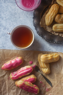 Homemade Strawberry Vanilla Eclairs With Pink Cream On Craft Paper And Vintage Plate And A Cup Of Black Tea On Light Background, Overhead Views. Delicious Breakfast, Traditional French Dessert