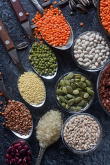 Raw organic cereal grains, seeds and beans (millet, rye,wheat, buckwheat, red and white beans, lentil, rice)  in wooden spoons and bowls on dark stone background. Healthy eating concept