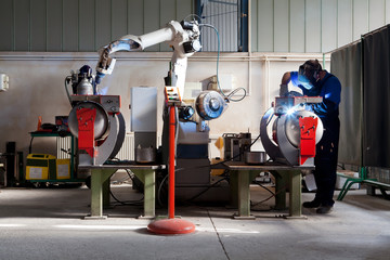 Man and robotic machine work together inside industrial building. The mechanical arm performs welds...