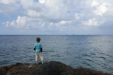 Fototapeta na wymiar A 6 year old boy in a shirt with a pensive look standing on a cliff, observing the calm sea and ships waiting in line for the port