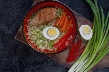 Ceramic bowl of traditional asian ramen soup with noodles, spring onion, chicken, sliced egg on dark stone table. Top view, selective focus.