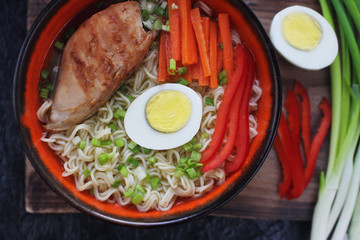 Ceramic bowl of traditional asian ramen soup with noodles, spring onion, chicken, sliced egg on dark stone table. Top view, selective focus.