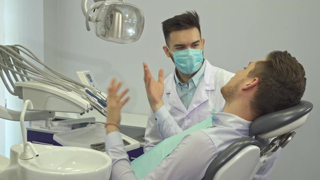 Handsome blond client showing thumb up at the dentist's office. Attractive caucasian patient approving dental specialist. Young man in the chair turning his face to the camera