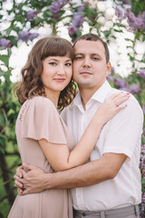Fototapeta premium beautiful young couple, the girl in beige light dress, the guy in the white shirt, cuddling, watching with a smile near the tree, greens, summer, lilac, close-up, nature