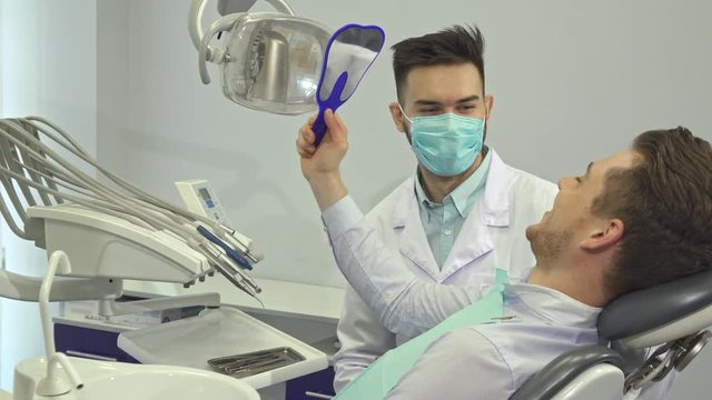 Attractive young patient looking at his teeth in the mirror at the dentist's office. Handsome blond man gesturing good job. Brunette caucasian doctor in mouth mask sitting on background of his client