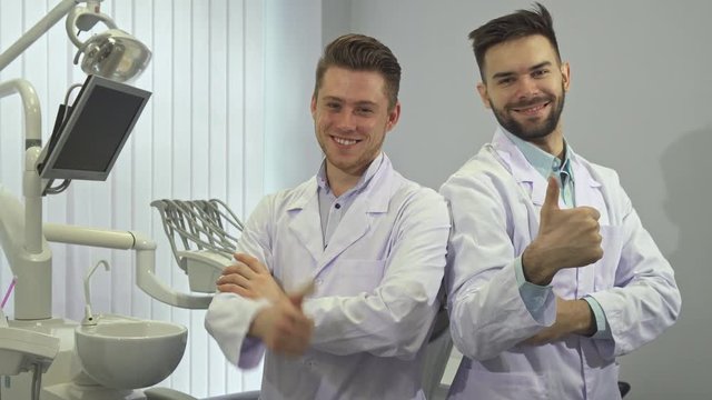 Two handsome caucasian dentists showing their thumbs up at the office. Attractive young doctors approving their work. Male dental specialists keeping their arms folded on the chests