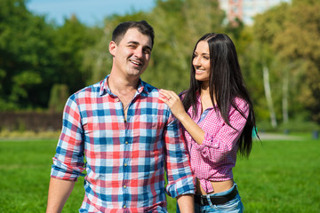 Fototapeta na wymiar Young beautiful loving couple in checked shirts and jeans standing on the green lawn and laughing. A girlfriend is holding her hand on her boyfriend’s shoulder. Outdoor shot