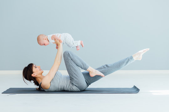 Sport, motherhood and active lifestyle concept - side view of young mother workout together with her baby over gray wall background. Mother having fun and playing with her little son.