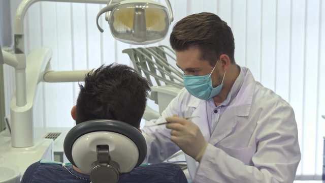 Young blond dentist examining his client's teeth at the office. Attractive caucasian doctor in mouth mask holding dental probe in his hand. Male medical worker bending over his patient