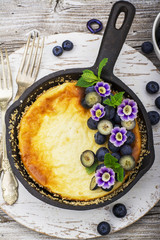 Healthy lunch: homemade cheesecake with blueberries and edible flowers in vintage black cast iron frying pan on wooden background. Top view