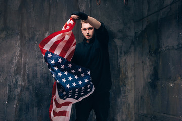 Young USA patriot with flying american flag. Strong man in black cloth on dark studio background....