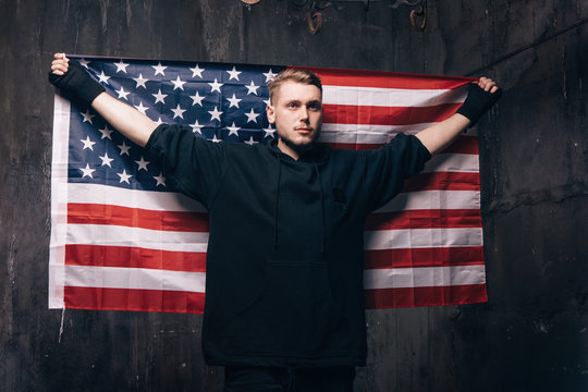Young man with USA national flag in studio. Strong guy in black cloth with stars and stripes behind him. Independence day, confidence, pride, fidelity to the nation, memorial day concept