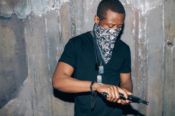 Black gang member checks his weapon. Gangster man with gun in hand on dark gray background. Outlaw, ghetto, murderer, armed attack concept - obrazy, fototapety, plakaty