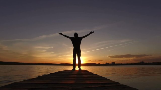 ilhouette of a guy on a dock for boats in the lake raises his hands up at sunset.