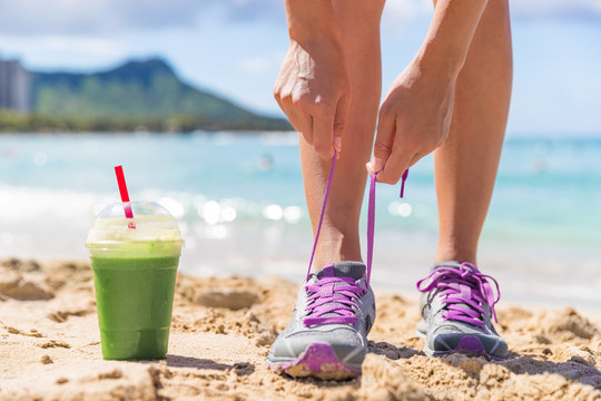 Green smoothie runner fitness woman tying running shoes at beach workout. Healthy lifestyle detox weight loss girl lacing trainers on Waikiki, Honolulu, Hawaii.