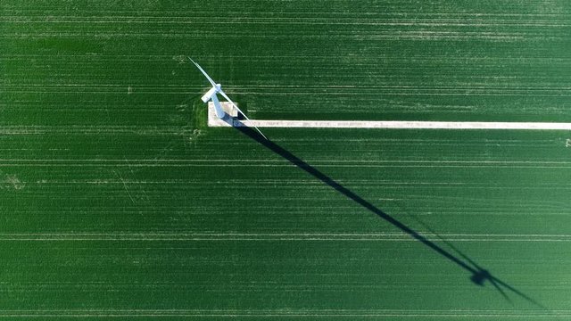 Aerial footage top-down view of wind turbine located on grass landscape blades turning fast producing sustainable energy also showing the maintenance road and wind turbines shadow on flat grass 4k