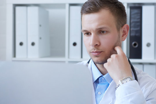 Young Male Doctor Using Computer On Desk In Clinic.