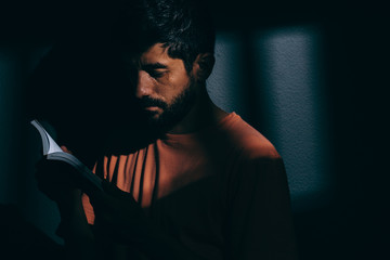 Prisoner man in dark cell reading a book or bible