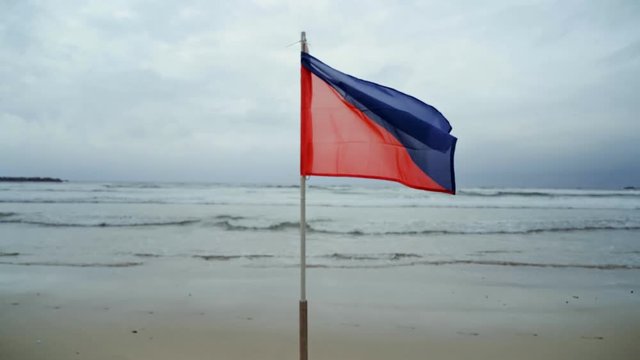 High medium hazard blue and red flag, waves and flaps in strong stormy winds in middle of grey and rainy beach, alarms surfers about dangerous waves, winds and marine life