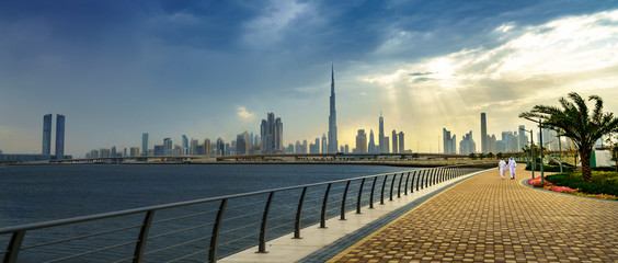 Panoramic view of business bay, downtown area of Dubai and two arab men take a walk on the promenade at cloudy day. UAE - obrazy, fototapety, plakaty