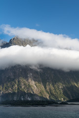 Fiordland National Park, New Zealand - March 16, 2017: Huge cliffs descend into the waters of Milford Sound under blue sky. Green forests at its base. White cloud band hangs midway the rock.