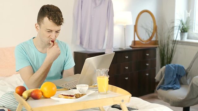 Young man eating breakfast in bed and using notebook, steadycam shot
