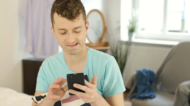 Young man sitting on bed and doing selfies on smartphone, steadycam shot

