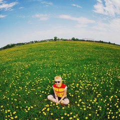 Portrait of young cute smiling blonde in a yellow and red waistcoat in sunglasses, sitting in a lawn among yellow flowers of dandelion. Beautiful skyline with fish-eye effect.