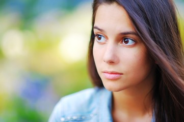 Fototapeta premium Young pretty brooding brown-eyed woman outdoors portrait. Soft sunny colors, blurred background. Close-up